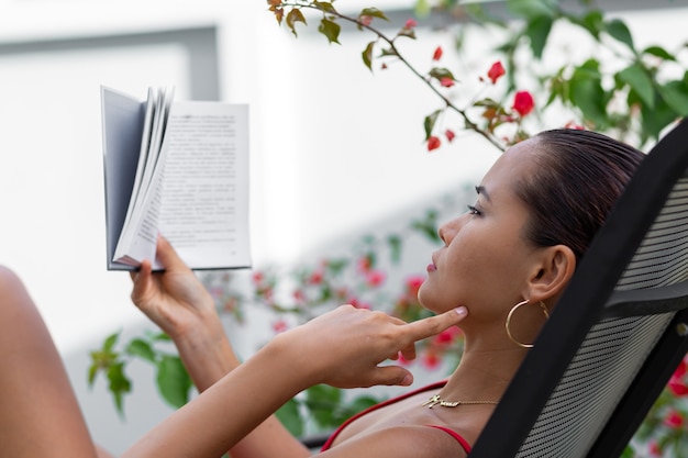 Free photo asian woman in swimsuit relax by pool on sunbed read book at villa by colorful flower tree