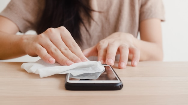 Asian woman using alcohol spray on tissue clean mobile phone before use it for protect coronavirus. Female clean surface for hygiene when social distancing stay at home and self quarantine time.