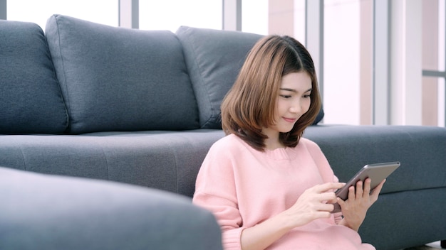 Asian woman using tablet while lying on home sofa in her living room. 