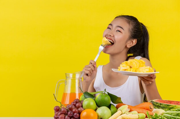 An Asian woman wearing a white tank top. Eating pineapple And the table is full of various kinds of fruits.