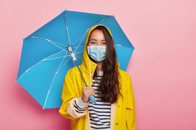 Free photo asian woman wears protective mask, faces air pollution during rainy day, stands under umbrella, dressed in yellow raincoat