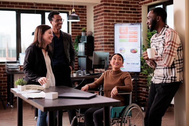 Asian woman in wheelchair and group of people laughing on job break, having fun in disability friendly office. Worker with chronic health condition chatting with cheerful colleagues.