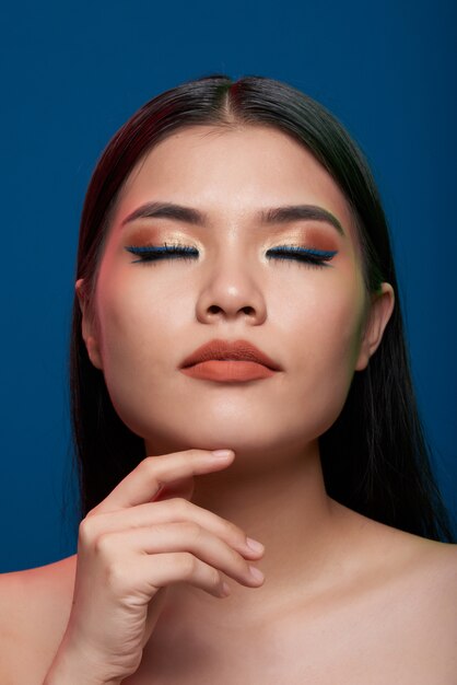 Asian woman with full makeup and bare shoulders posing with closed eyes and finger on chin