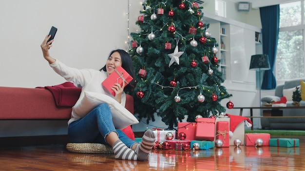 Asian women celebrate Christmas festival. Female teen relax happy holding Gift and using smartphone selfie with Christmas tree enjoy xmas winter holidays in living room at home.