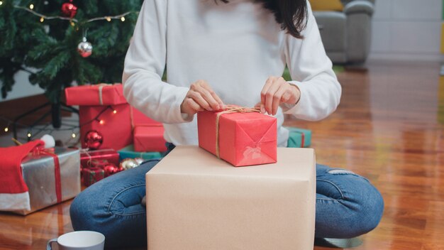 Asian women celebrate Christmas festival. Female teen wear sweater and Christmas hat relax happy wrapping gifts near Christmas tree enjoy xmas winter holidays together in living room at home.