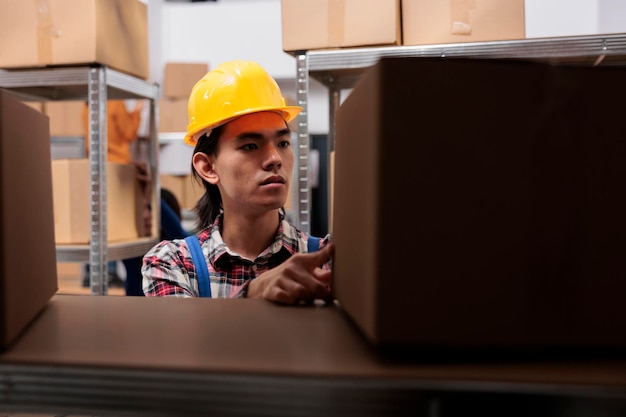 Free photo asian young postal worker doing parcels organization in warehouse. storehouse order picker in yellow hardhat looking at packages on shelf and searching for cardboard box close up