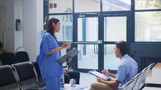 Free Photo assistant standing at hospital counter desk, writing medical expertise on clipboard. asian patient calls the nurse to help him with insurance information during checkup visit in waiting room