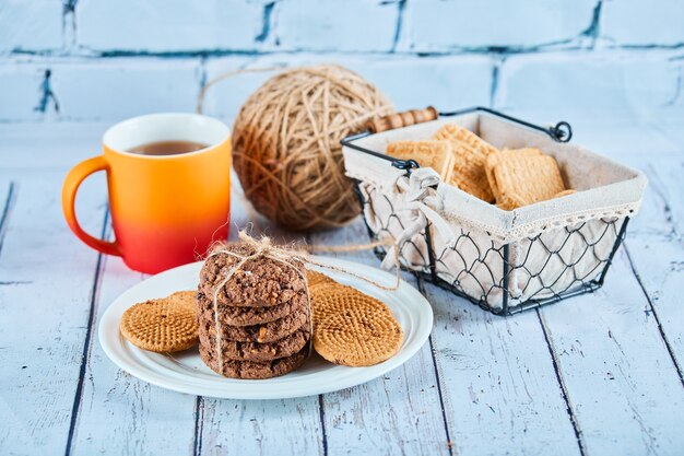 Assorted biscuits in plate and basket and a cup on blue table.