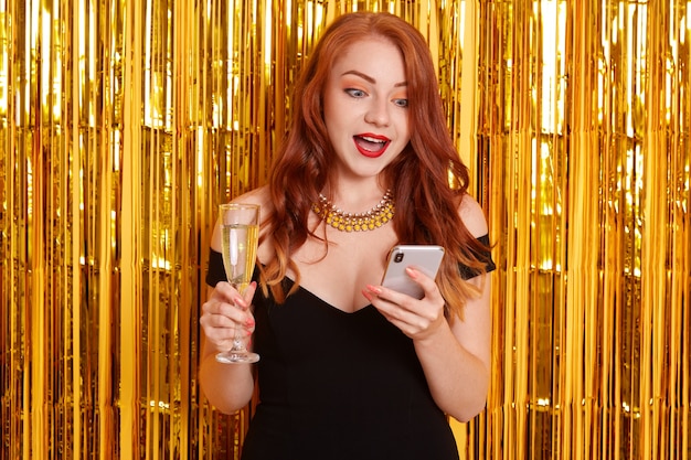 Free photo astonished female with red lips and widely opened mouth, looking with shocked facial expression at her device, girl wearing black dress, posing isolated over wall decorated with golden tinsel.