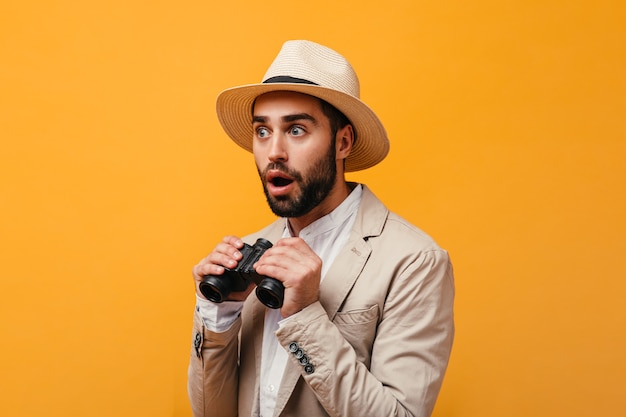 Astonished guy in hat holding binoculars on orange wall