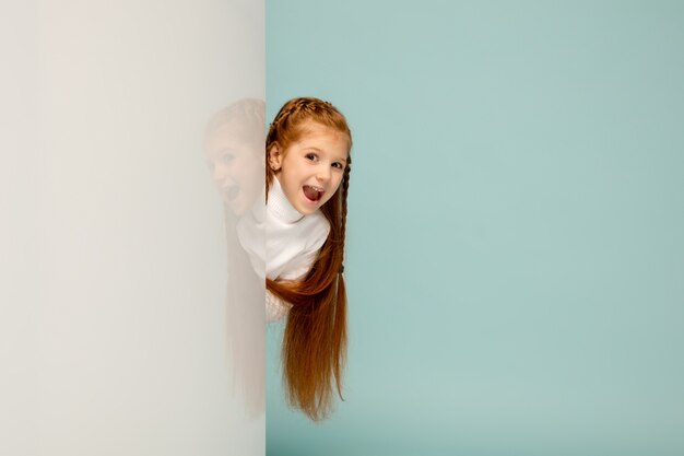 Astonished. Happy kid, girl isolated on blue studio background. Looks happy, cheerful. Copyspace for ad. Childhood, education, emotions, facial expression concept. Peeking out from behind the wall.