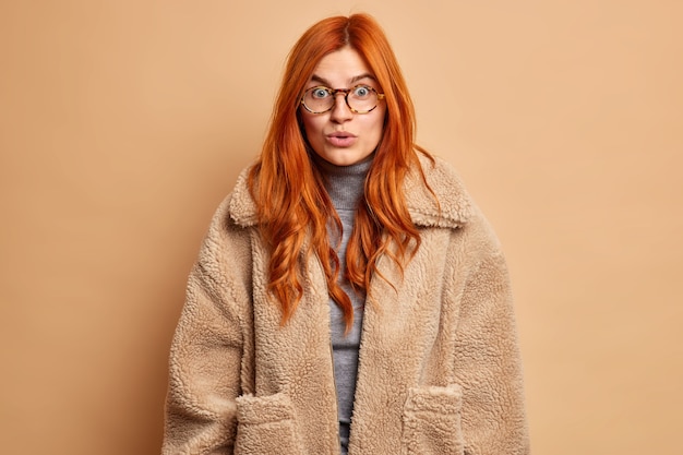 Astonished redhead European woman hears incredible rumors or secret impressed by something wears transparent glasses and fur brown coat.