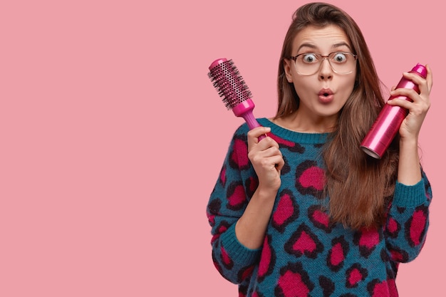 Astonished young lady has mess on head, prepares for special occasion, makes coiffure, surprised to look at herself in mirror