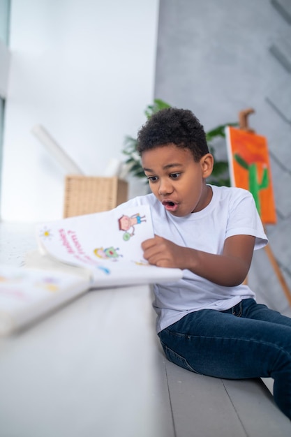 Free photo astonishment. dark-skinned school-age boy in white tshirt looking in surprise at book with open mouth sitting in bright room in daylight