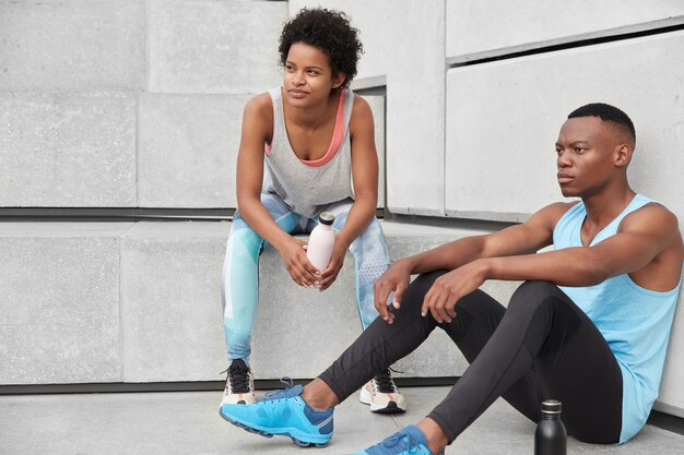 Athlete female and male runner sit at stairs, being deep in thoughts, dressed in casual clothes, drink coffee from sport clothes, feel tired rest after jogging. People, motivation, fitness concept