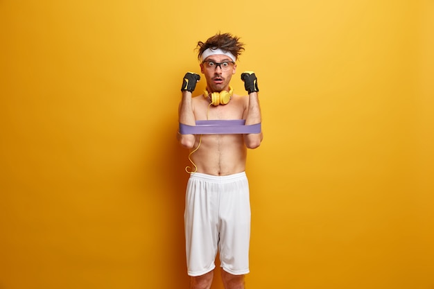 Free photo athlete man develops hand muscles with fitness gum, stares with shocked expression, wears white shorts, headband, uses stereo headphones, isolated on yellow wall. stay strong and healthy