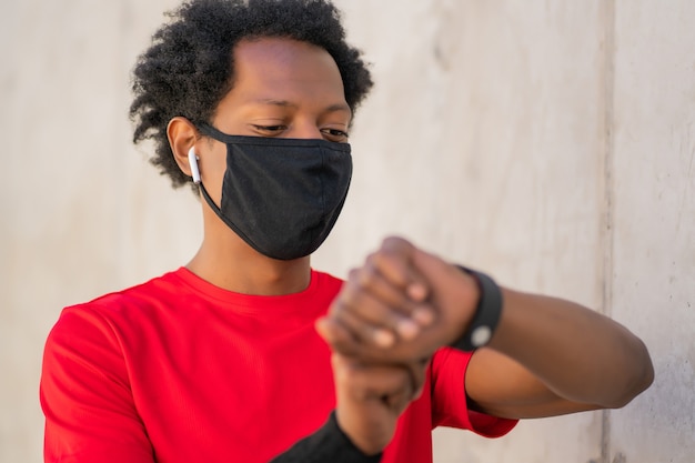 Athletic man wearing face mask and checking time on his smart watch while work out outdoors. New normal lifestyle. Sport and healthy lifestyle concept.