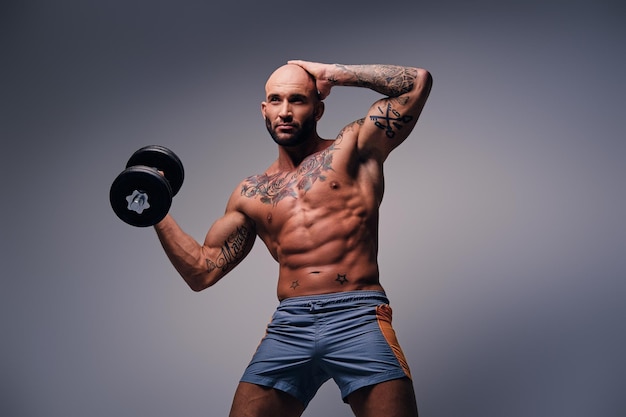 Athletic shaved head male with tattoos on his torso and arms holds dumbbell isolated on grey vignette background.