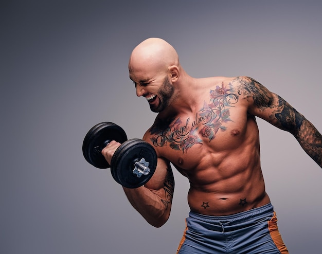 Athletic shaved head male with tattoos on his torso and arms holds dumbbell isolated on grey vignette background.