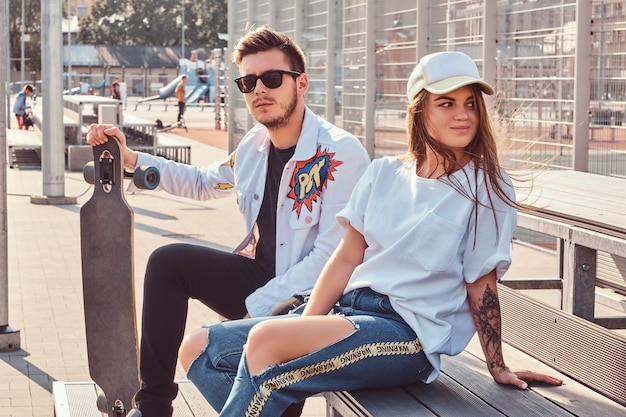 Free photo atractive couple of trendy dressed young hipsters with skateboards sitting on bench at city sports complex on sunny day, with warm toned.