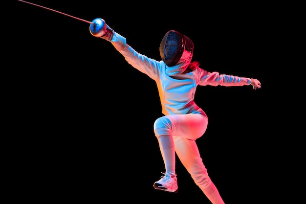 Free photo attacking. teen girl in fencing costume with sword in hand isolated on black background, neon light. young model practicing and training in motion, action. copyspace. sport, youth, healthy lifestyle.