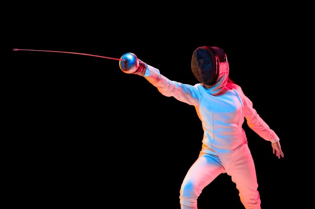 Attacking. Teen girl in fencing costume with sword in hand isolated on black wall, neon light. Young model practicing and training in motion, action. Copyspace. Sport, youth, healthy lifestyle.