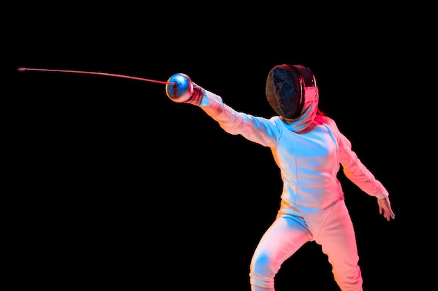 Attacking. Teen girl in fencing costume with sword in hand isolated on black wall, neon light. Young model practicing and training in motion, action. Copyspace. Sport, youth, healthy lifestyle.