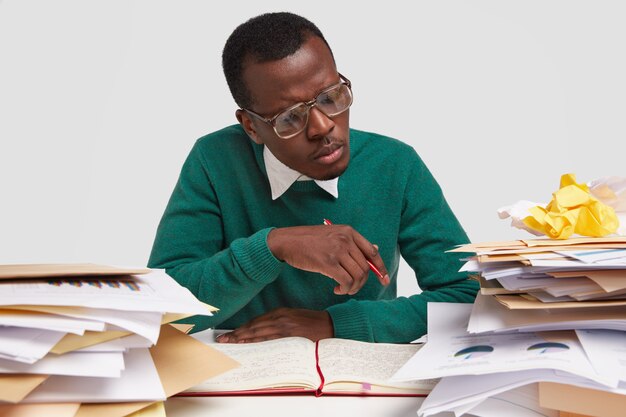 Attentive dark skinned man in big eyewear, looks seriously at pie chart, writes report after studying documentation, dressed in green jumper, isolated over white background. People