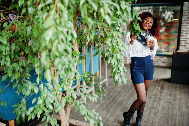 Attractive african american curly girl in white blouse and blue shorts posed at cafe against tree with latte at hand