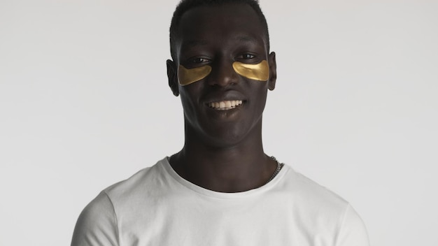 Attractive African American guy with gold eye patches smiling on camera over white background. Facial treatment