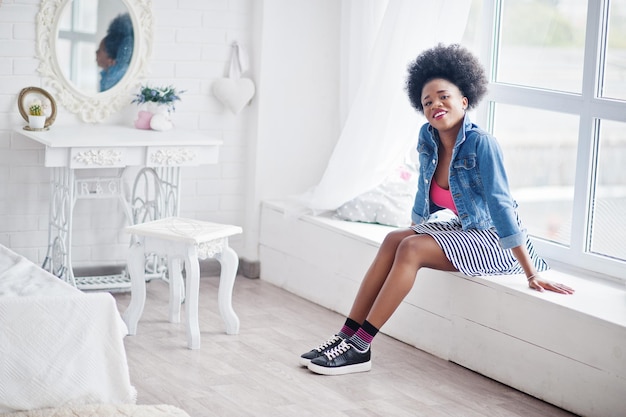 Attractive african american woman with afro hair wear on skirt and jeans jacket posed at white room Fashionable black model