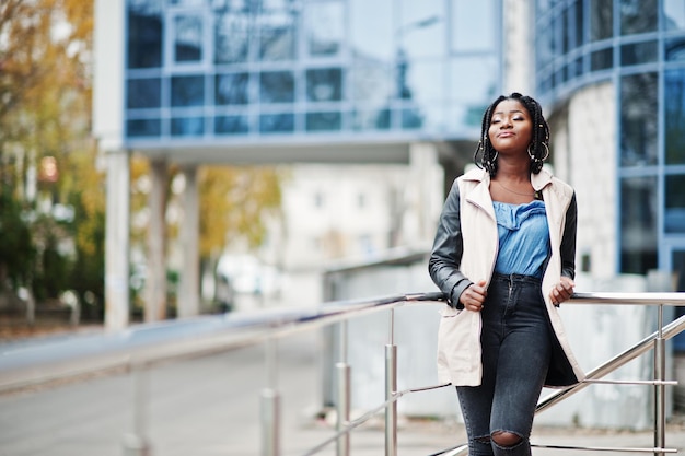 Attractive african american woman with dreads in jacket posed near railings against modern multistory building