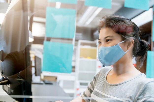 Attractive asian female navy blue sweater working while wearing protective virus mask working in co woking space that have plastic partition social distancing for seat new normal lifestyle