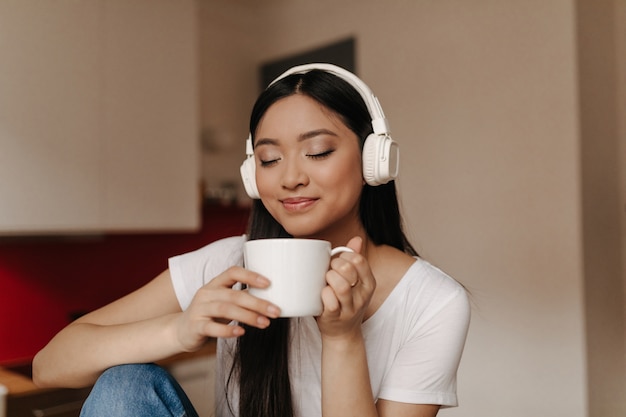 Free Photo attractive asian woman in headphones inhale aroma of tea, holding cup and smiling