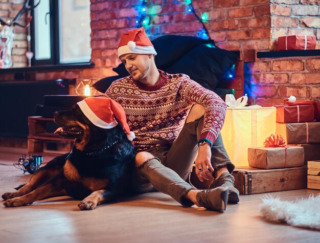 Attractive bearded hipster male sits on a floor with his Rottweiler dog in a room with Christmas decoration.