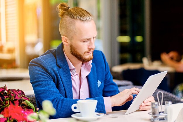 Attractive bearded redhead male in a pink shirt and blue jacket sits at the table in a cafe and using a tablet PC.