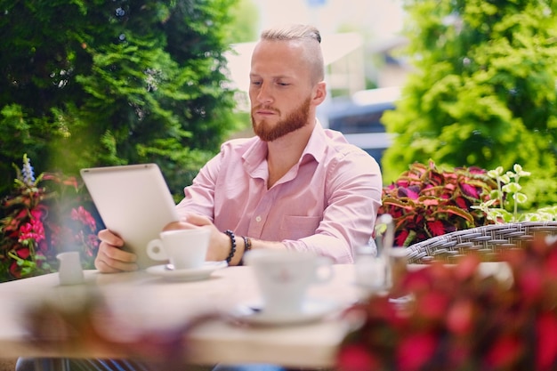 Free Photo attractive bearded redhead male in a pink shirt sits at the table in a cafe and using a tablet pc.