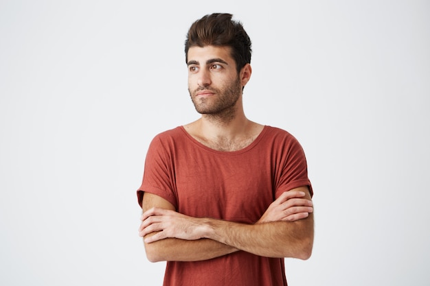 Attractive bearded sun-tanned man having pleased look standing crossed hands against white wall. Student wearing casual t-shirt shirt waiting for his girlfriend