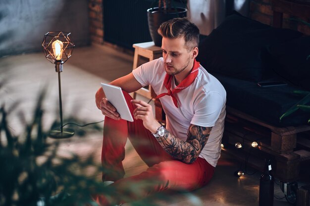 Attractive blond male with tattoos on arms, dressed in a red Christmas pants using tablet PC in a room with loft interior.