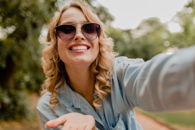 Free photo attractive blond smiling woman walking in park in summer outfit taking selfie photo on phone