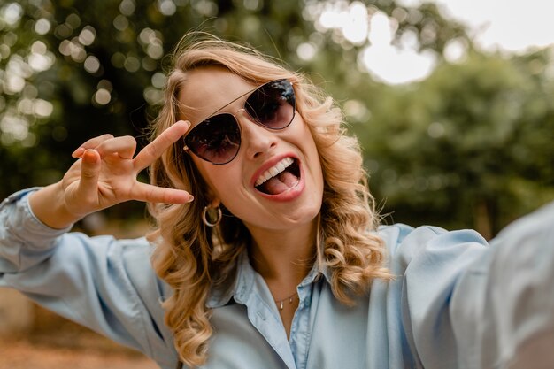 Attractive blond smiling woman walking in park in summer outfit taking selfie photo on phone