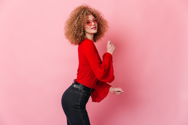 Attractive blonde girl in sunglasses dressed in top with wide sleeves and dark pants dancing on pink space.