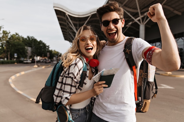 Free photo attractive blonde woman in sunglasses and man in white tee smiles and takes selfie near airport