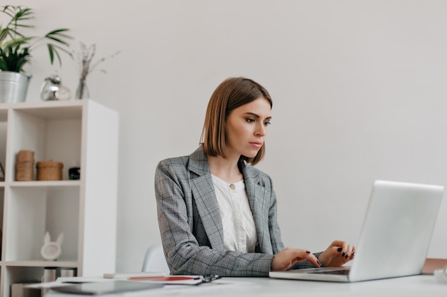 Attractive blonde woman typing letter in laptop at her workplace. portrait of lady in stylish jacket in bright office.