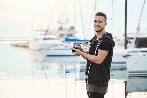 Attractive boyfriend focused on his hobby during walk with girlfriend. Portrait of guy standing in harbour near yachts, holding camera, looking aside while searching for great shot.