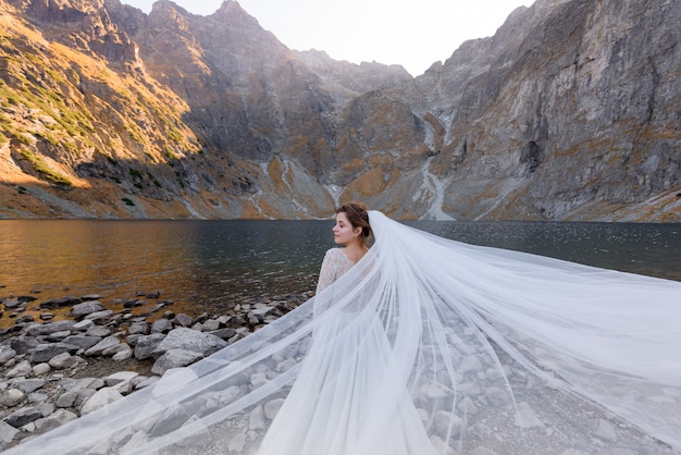 Free Photo attractive bride with closed eyes and wavy veil is standing in front of lake surrounded with autumn mountains on the sunny day