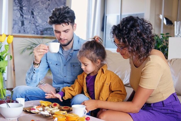 Free photo attractive brunette female in eyeglasses, bearded stylish male and their cute little daughter sits on a couch and eat lunch in a living room.