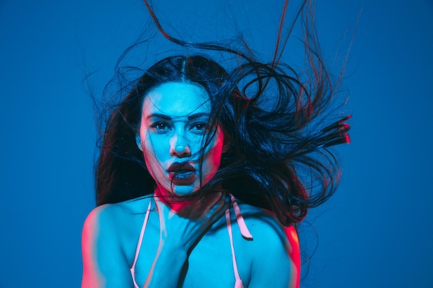 Attractive brunette model on blue  wall in neon light. Beautiful women in underwear posing with flying hair and dark make up. Concept of sensuality, style, fashion industry, characters.