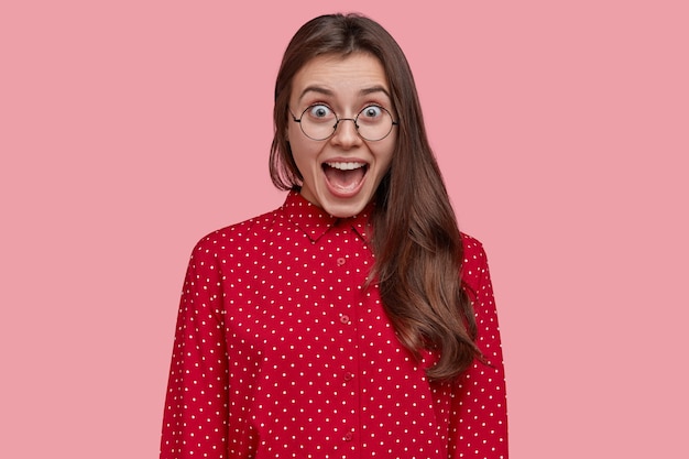 Attractive brunette overjoyed woman opens mouth and yells from joy, wears pink polka dot blouse, spectacles, glad with her success