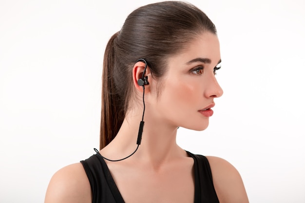 Attractive brunette woman in jogging black top listening to music on earphones posing isolated on white wall ponytail hairstyle